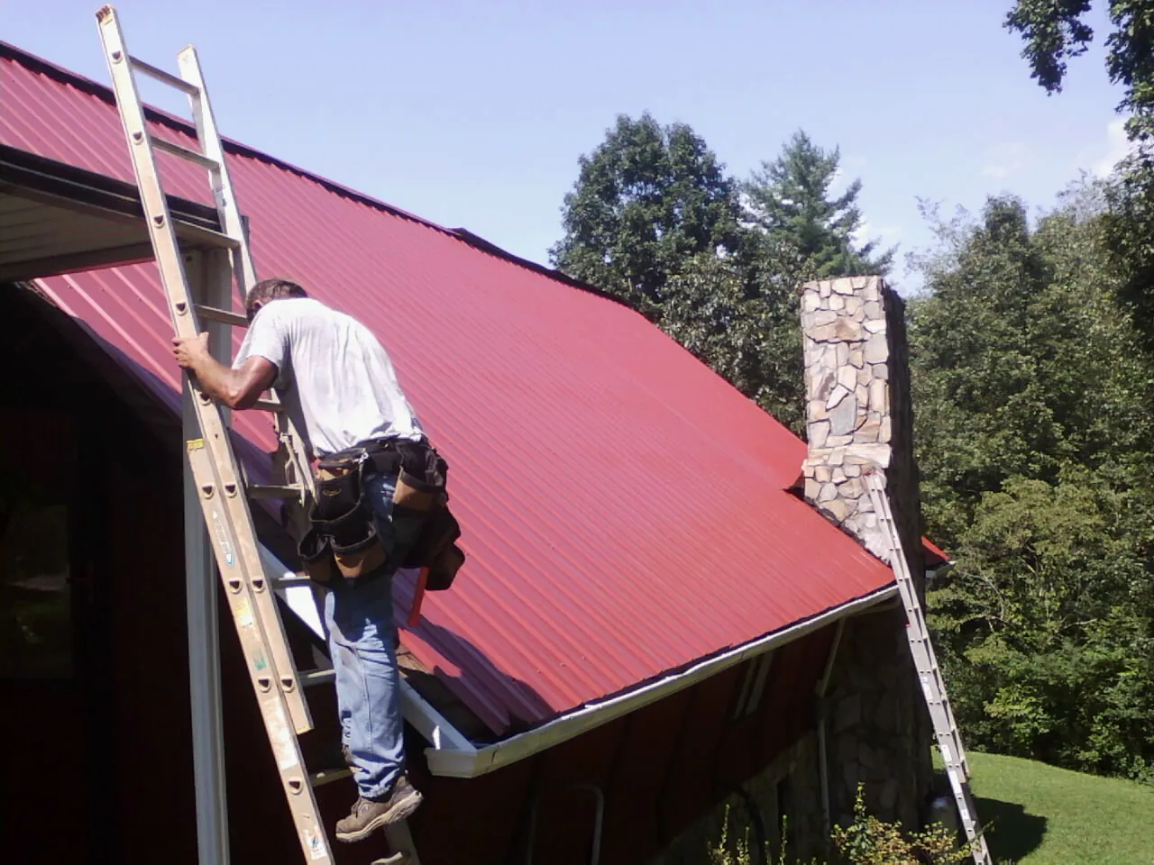 Roofer on a ladder beside a red metal roof and stone chimney — Soffit & Fascia Repair by Roofing Brothers in Central City